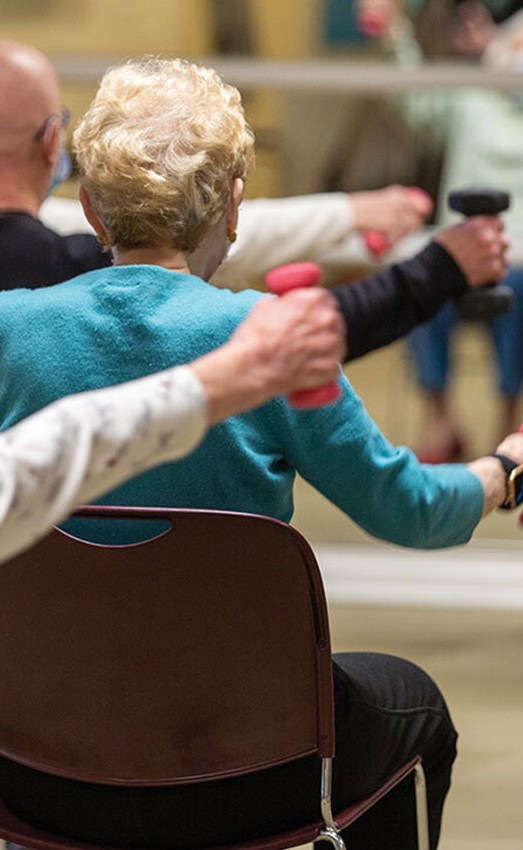 Residents in an exercise class sitting in chair lifting waits facing a mirror.