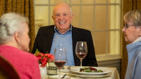 Three residents sit at a dining table. A man looks at the camera smiling with a glass of wine.