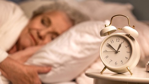 An alarm clock on a nightstand with a woman slipping in the background.